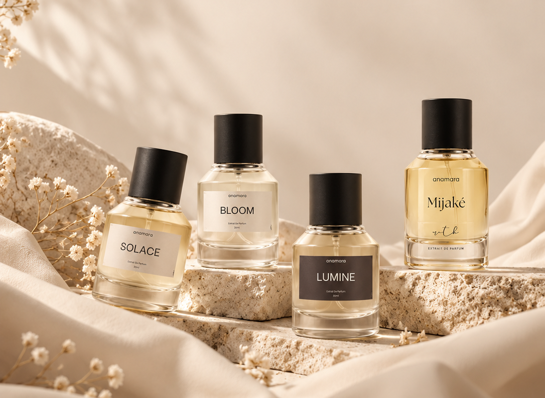 Four perfume bottles from the brand anmr., including Solace, Bloom, Lumine, and Mijaké, arranged on a textured stone slab with draped cream fabric and dried flowers.